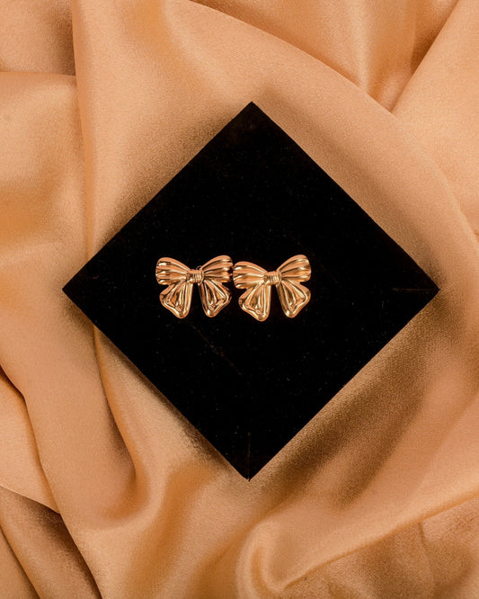 Pair of gold bow-shaped earrings on a black box with beige fabric background