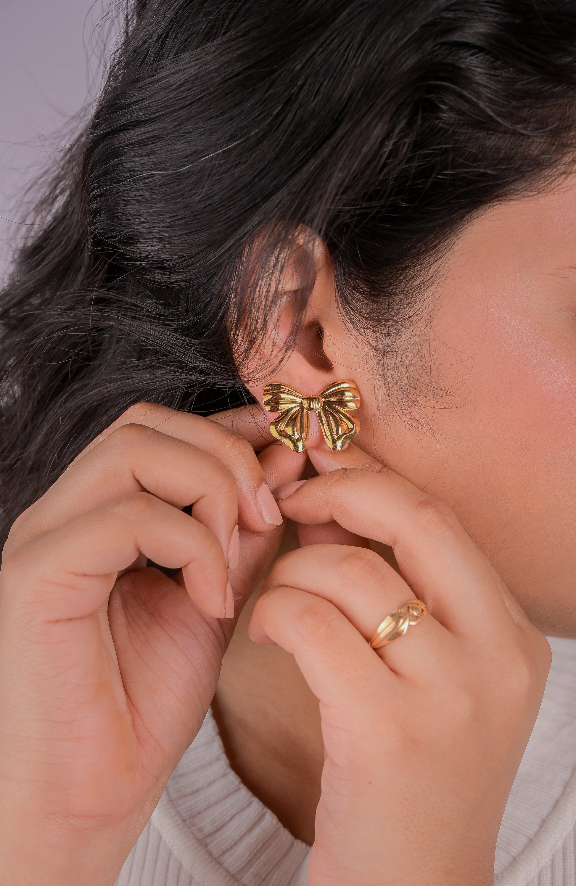Person wearing gold bow-shaped earrings and a ring, with a neutral background